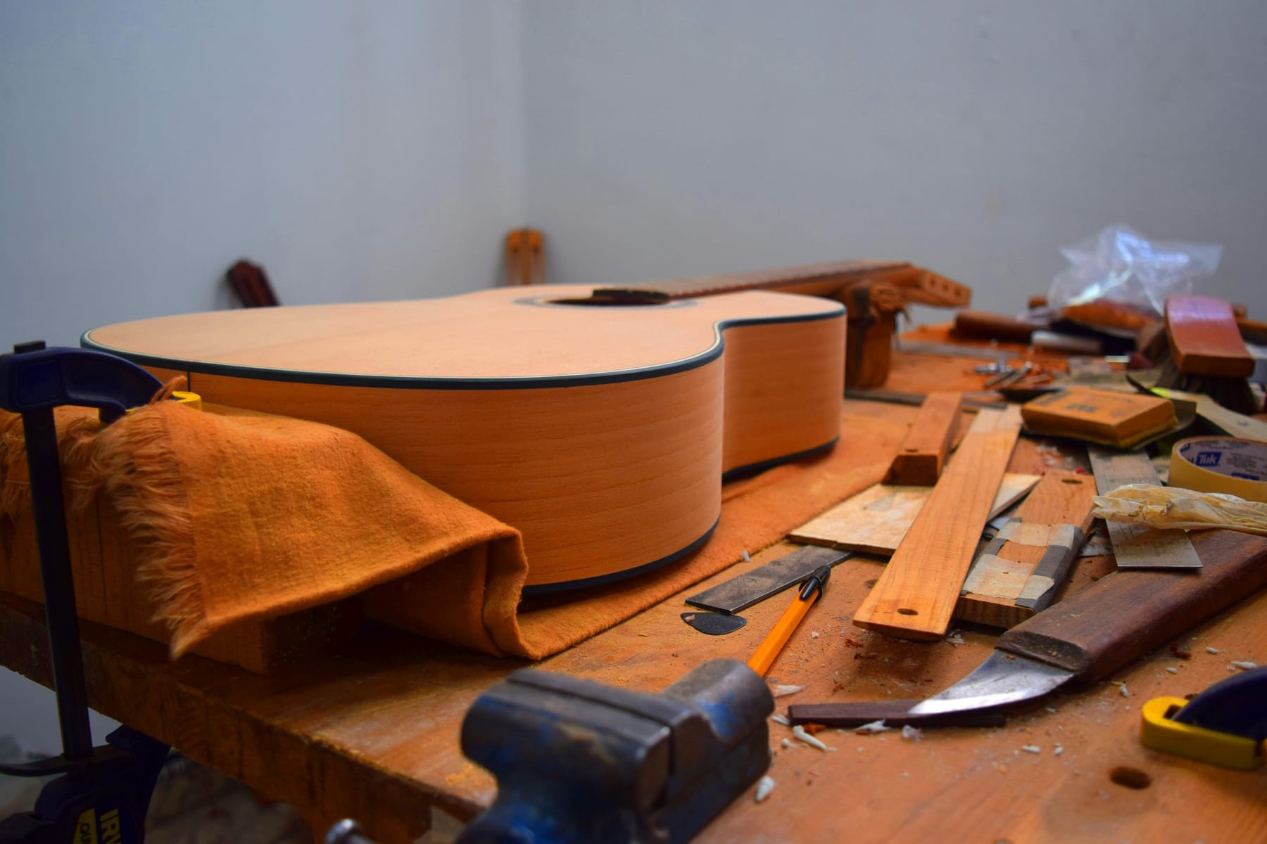 An unfinished acoustic guitar body upright on a workbench, beside a mallet, a chisel and offcuts of wood.