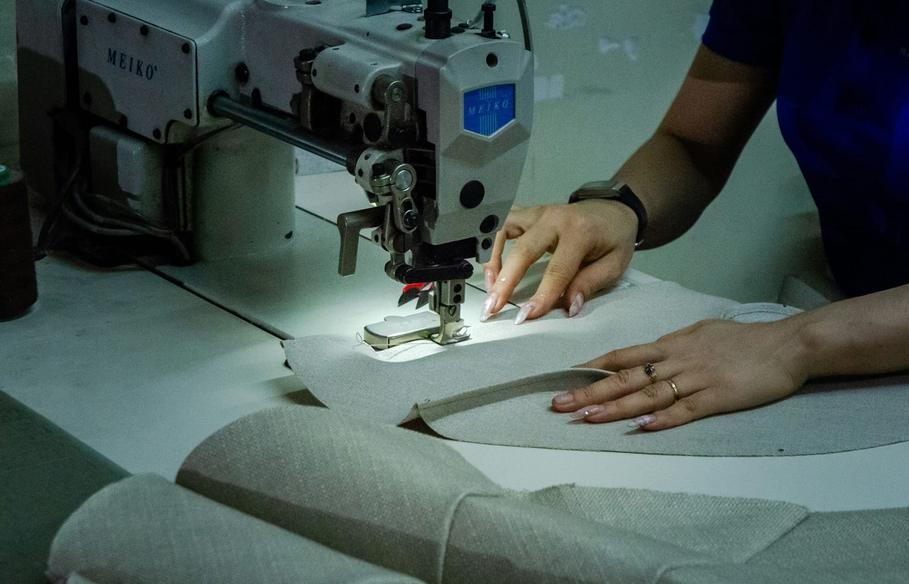 Hands guiding a panel of pale cloth through an industrial sewing machine.