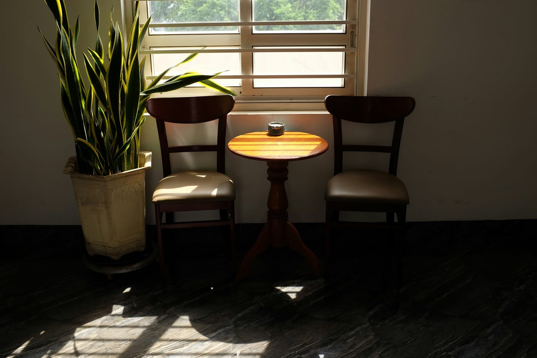 Two plain wooden chairs either side of a small round table, by a bright window with a plant beside it.