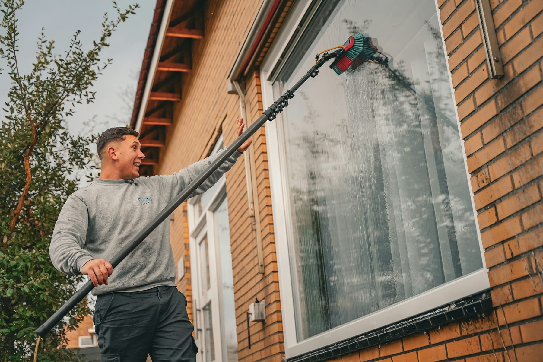 A window cleaner standing on the ground, reaching an upper-storey window with a long pole.