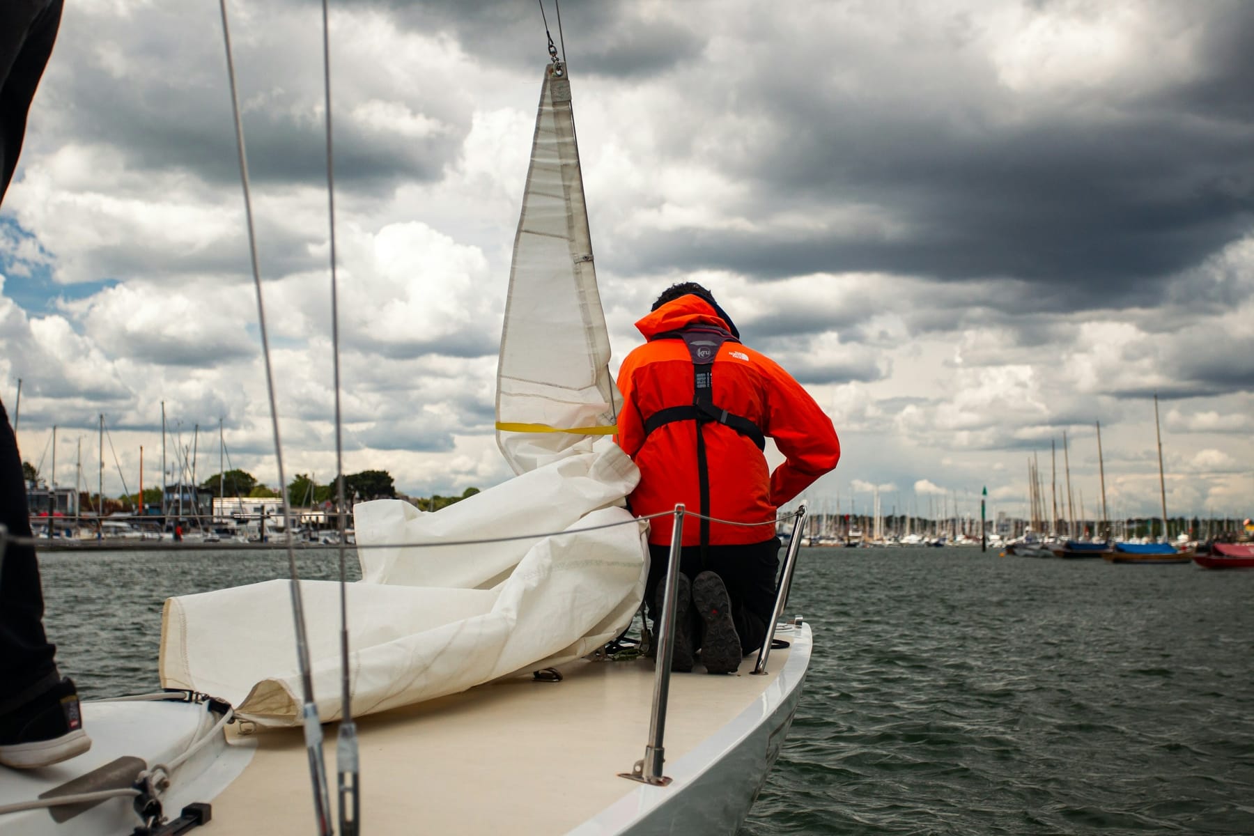 A sailor in a lifejacket kneeling on the foredeck, flaking a sail under a grey overcast sky.