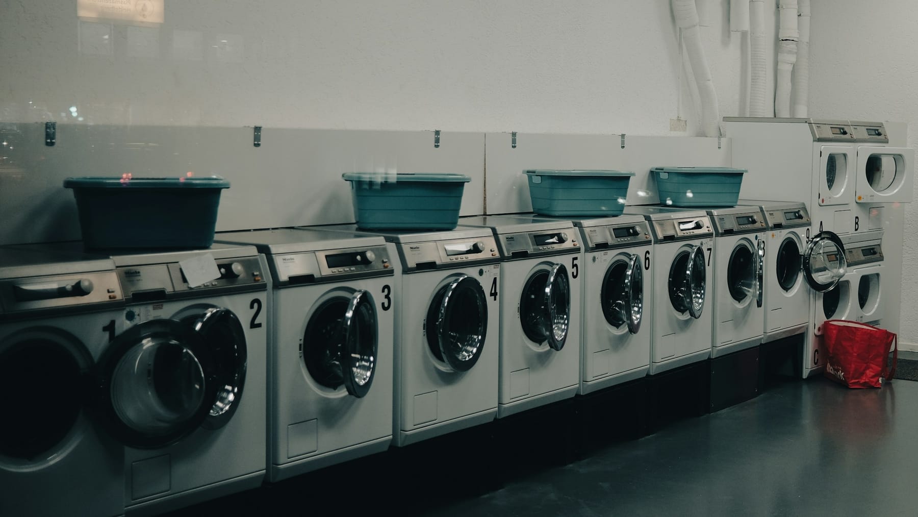 A row of numbered front-loading washers along a wall, with stacked plastic baskets on top of them.
