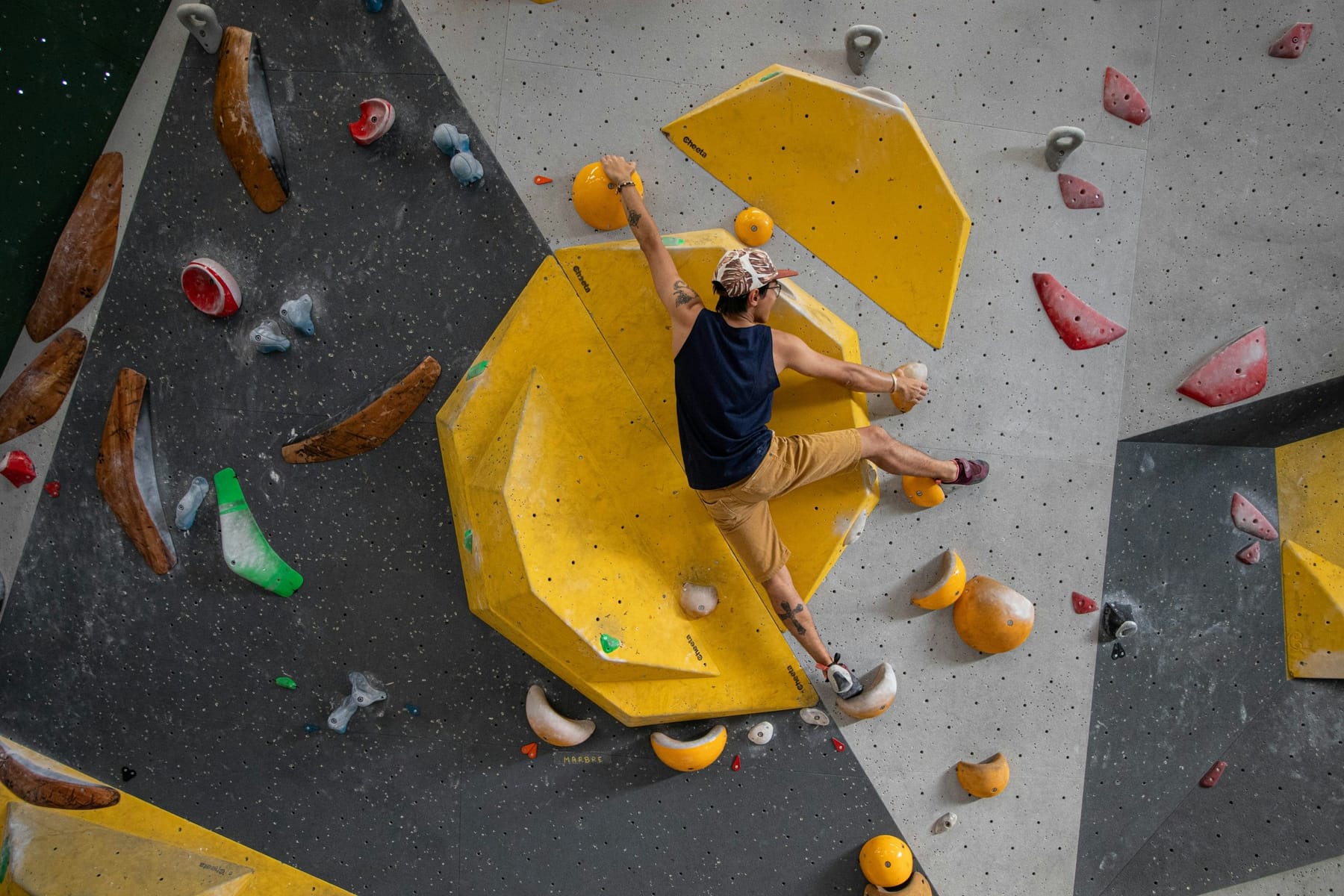 A climber part-way up a bouldering wall, pulling on a large yellow volume, with matting below and no rope of any kind.