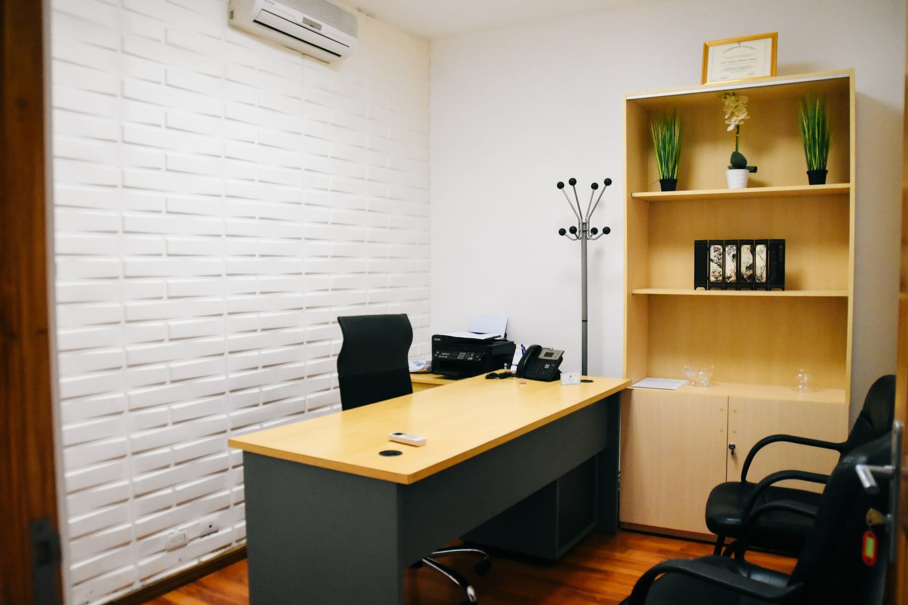 A plain small office with a desk, an office chair, a bookshelf and a coat stand.