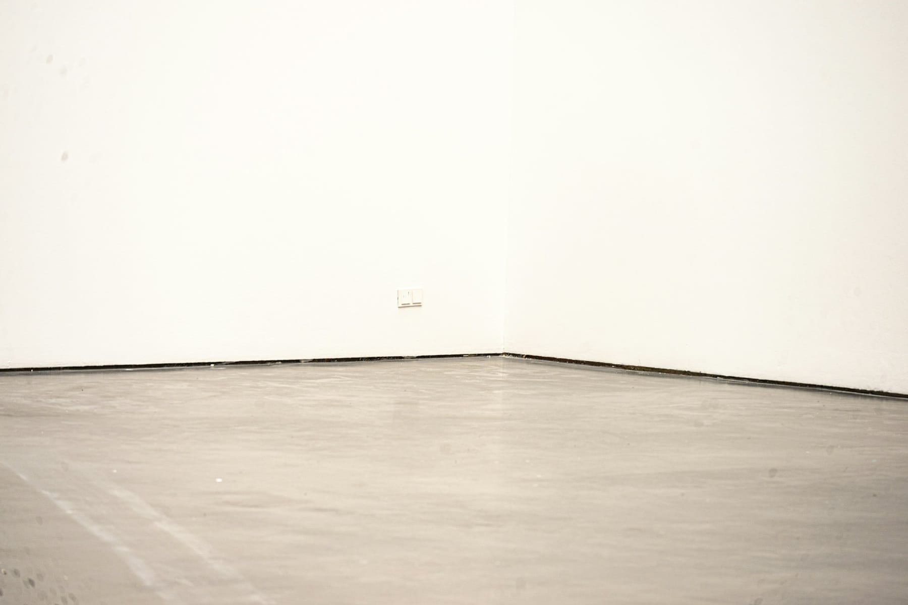 An empty white gallery corner meeting a bare concrete floor, no work hung.