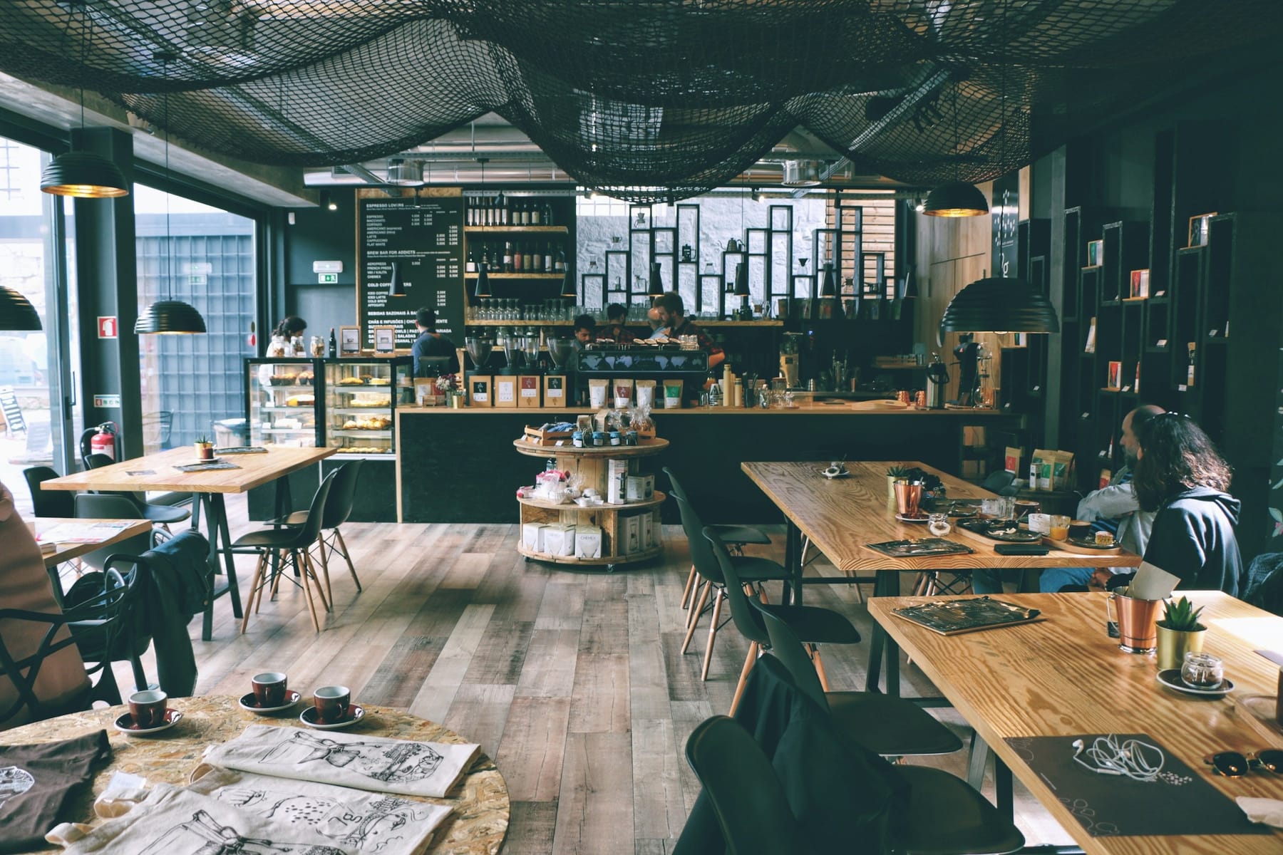 A café interior with long shared tables, a service counter at the back, and daylight across a wooden floor.