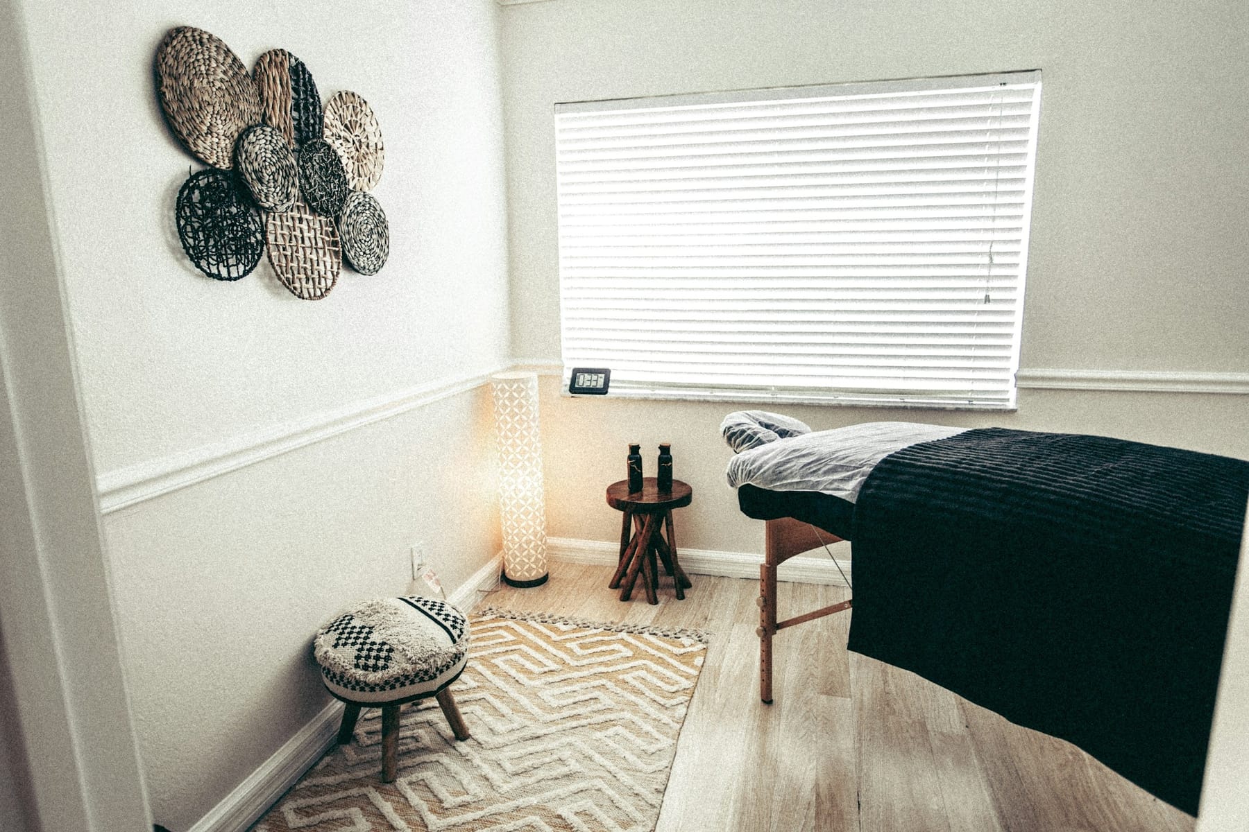 An empty treatment room: a covered table under a shuttered window, a floor lamp, a small stool holding two bottles, and a patterned rug.