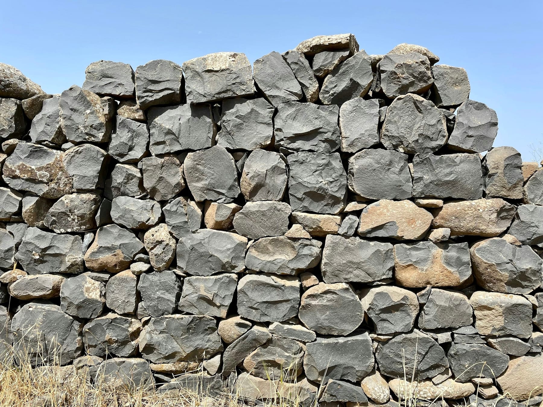 A dry-stacked wall of angular grey basalt, three courses visible, with dry grass growing at its base and open sky above.