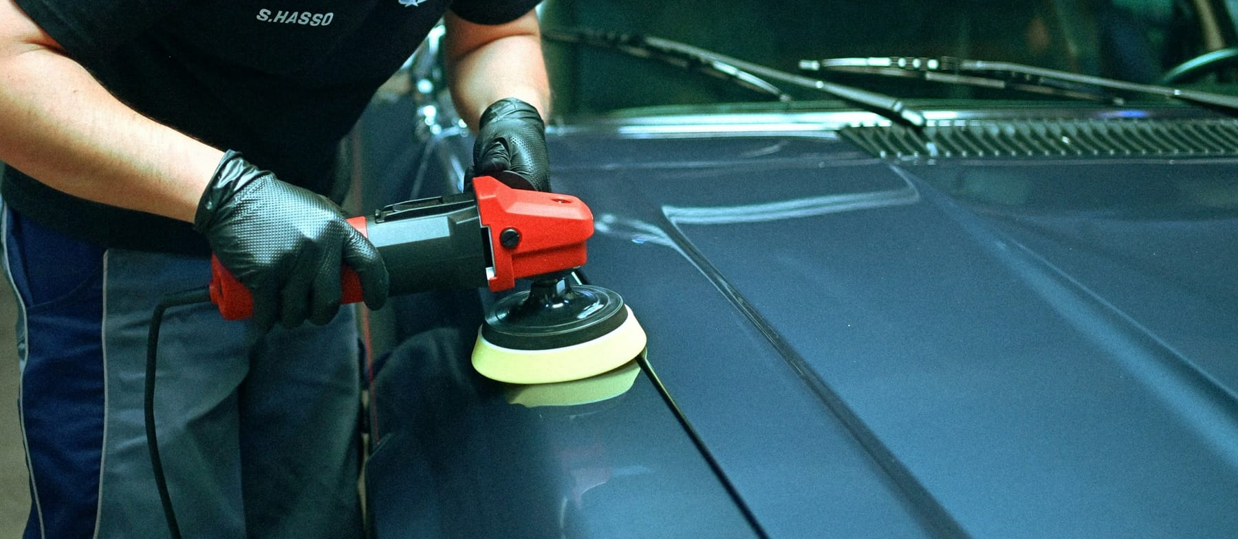 A dual-action polisher with a yellow pad working across a dark blue car panel.