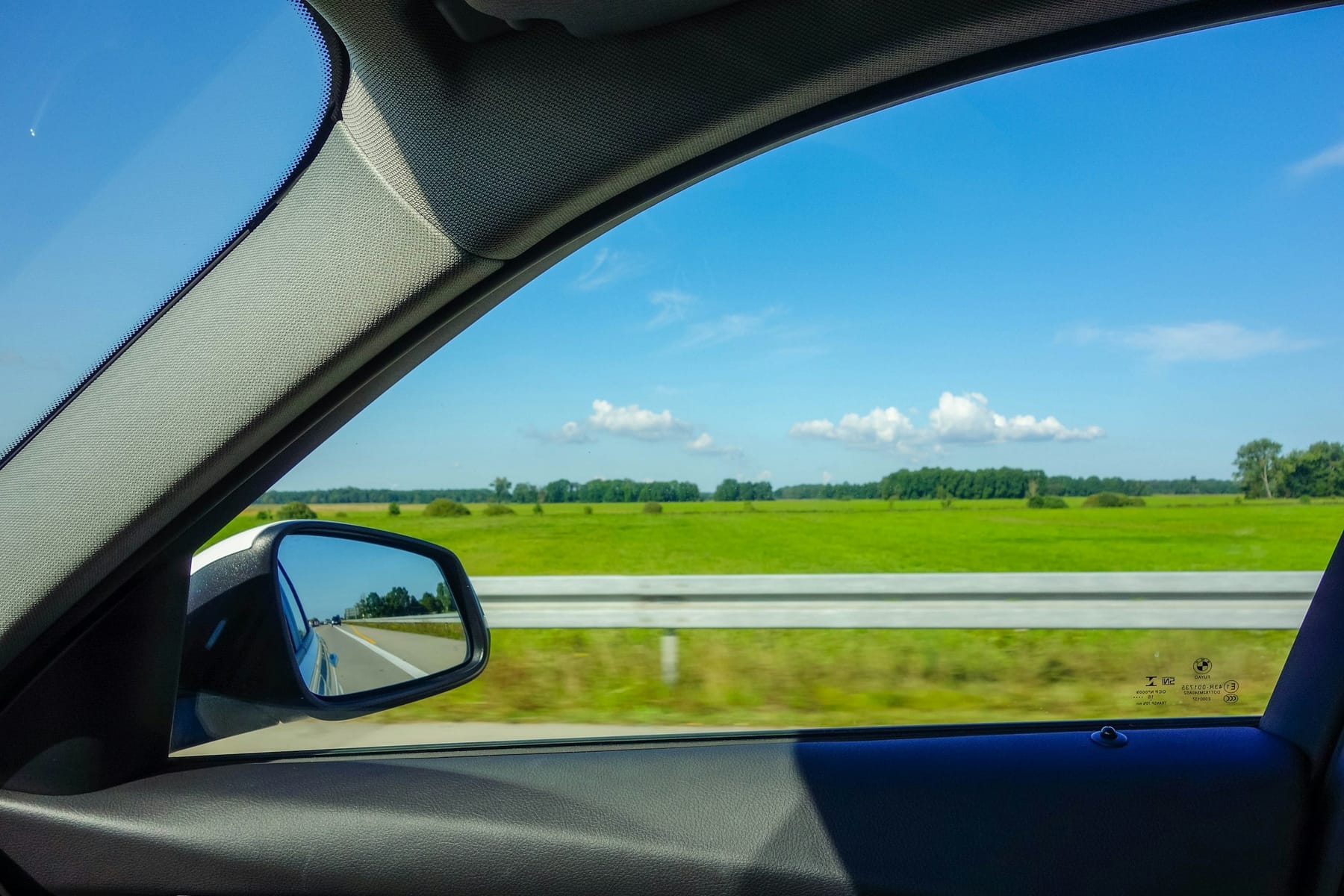 The view out of a car's side window in daylight: a green field, a guard rail and blue sky.