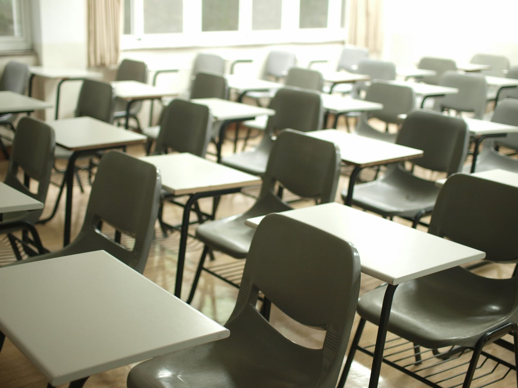 Rows of plain tables and grey chairs in an empty classroom, lit from a tall window at the back.