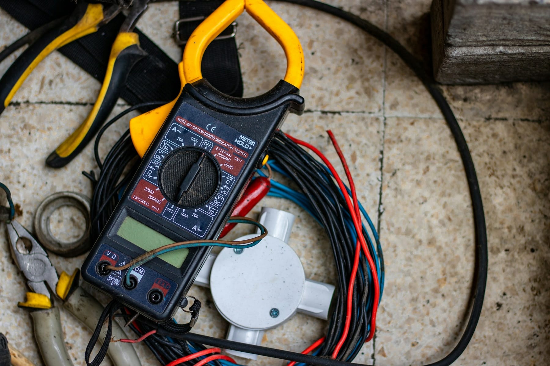 A clamp meter lying on a tiled floor beside a coiled bundle of red, black and blue wire and an open junction box.