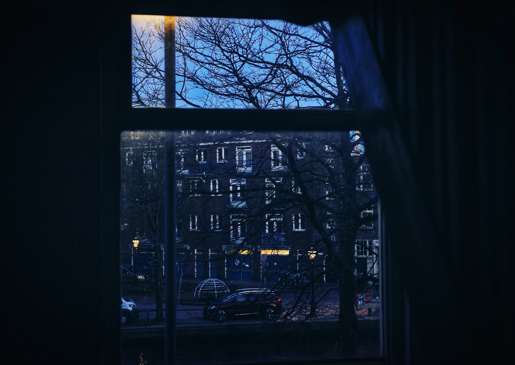 A window at dusk seen from inside a dark room, looking out at a building and bare trees.