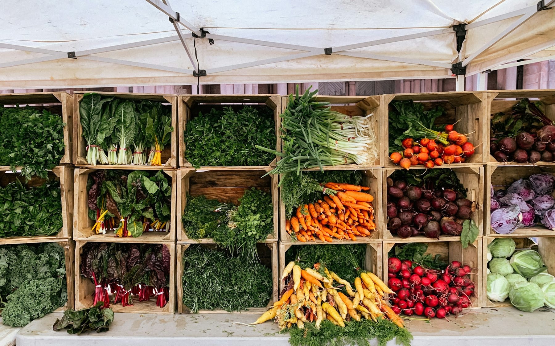 Wooden crates in a market stall holding greens, bunched carrots, beetroot, and squash blossoms, in flat daylight.